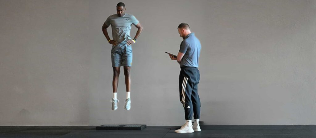 Plyometrisches Training in der späten Reha: Ein Athlet führt einen Sprung über einen Vault aus, während ein Coach Technik und Landung überwacht.