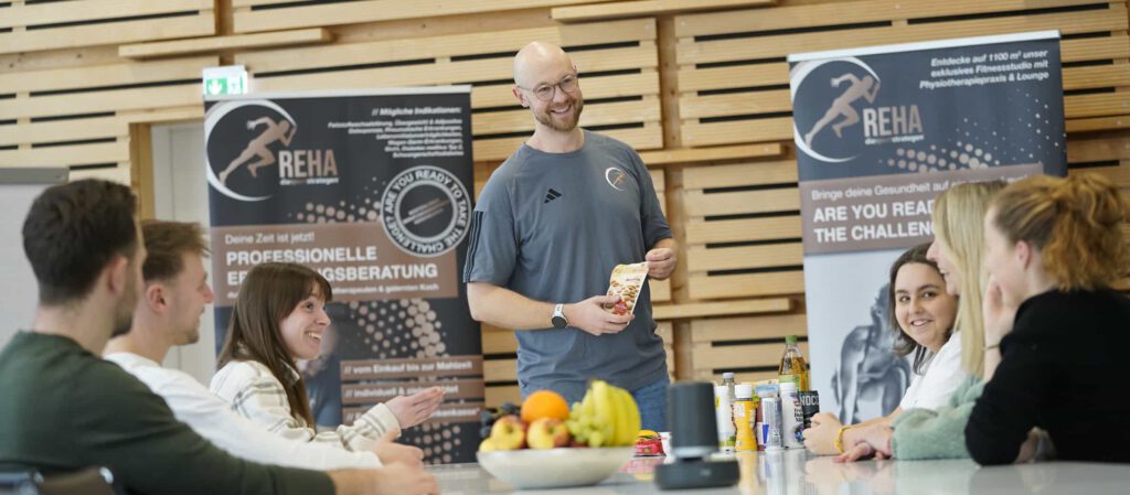 Betriebliche Gesundheitsförderung – Ernährungstherapeut Sven steht lächelnd vor einer Gruppe von Menschen. Im Hintergrund ist eine Präsentationsfläche mit der Aufschrift "Professionelle Ernährungsberatung" zu sehen. Der Raum ist hell und modern, die Teilnehmenden sitzen an einem großen Tisch und schauen aufmerksam zu ihm. Die Stimmung wirkt locker und informativ.