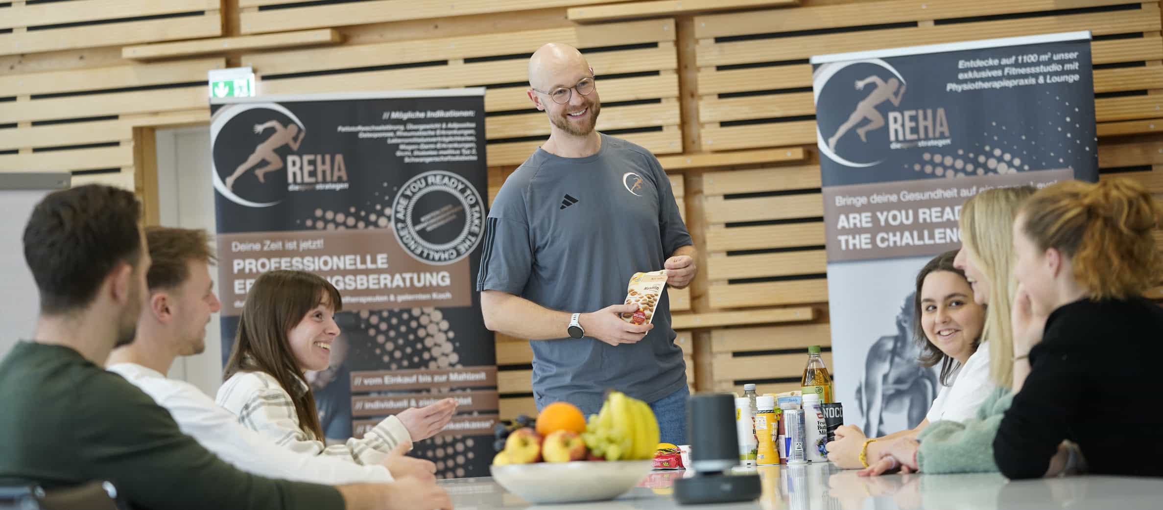 Betriebliche Gesundheitsförderung – Ernährungstherapeut Sven steht lächelnd vor einer Gruppe von Menschen. Im Hintergrund ist eine Präsentationsfläche mit der Aufschrift "Professionelle Ernährungsberatung" zu sehen. Der Raum ist hell und modern, die Teilnehmenden sitzen an einem großen Tisch und schauen aufmerksam zu ihm. Die Stimmung wirkt locker und informativ.