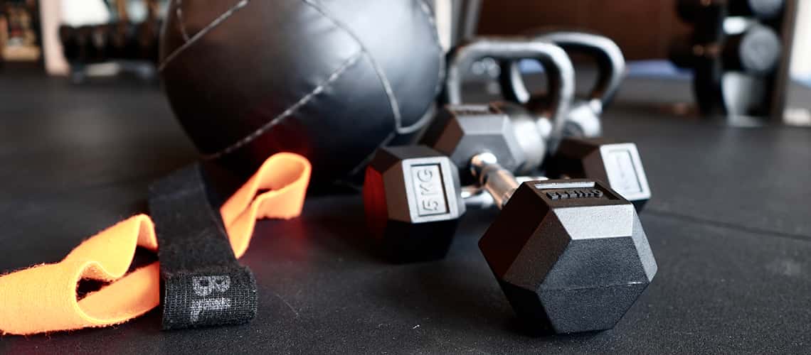 Hanteln, ein Theraband und ein Medizinball liegen auf dem Boden in einem modernen Fitnessstudio und symbolisieren vielseitiges Krafttraining für die Gesundheit.