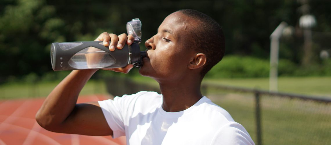 Sporternährung im Sommer: Mann trinkt nach dem Training auf dem Sportplatz Wasser aus der Sportflasche zur optimalen Hydration.